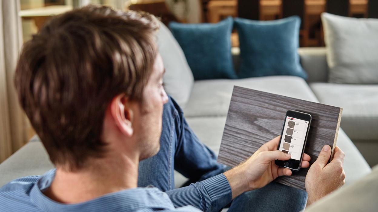 Holzdekor-Vergleich Junger Mann auf Sofa vergleicht Holzoberflächen mit dem Handy